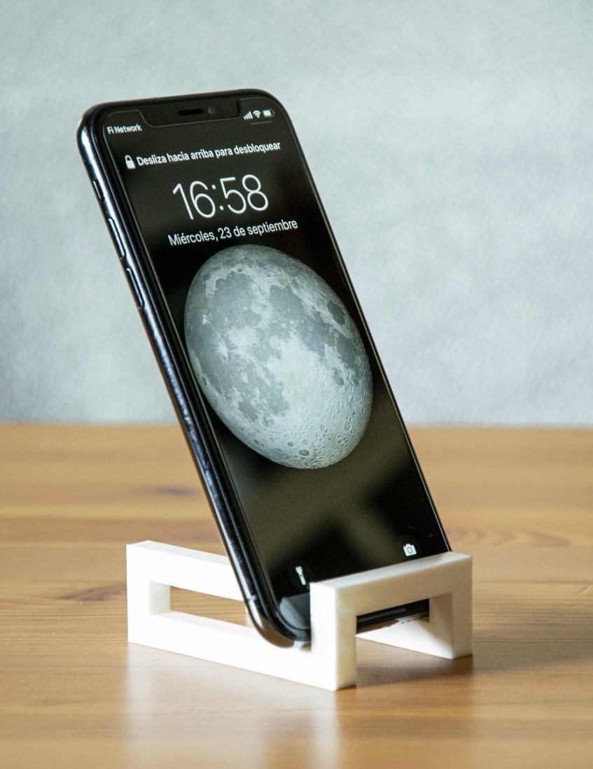 Smartphone with moon wallpaper on screen, placed on a white stand on a wooden surface.