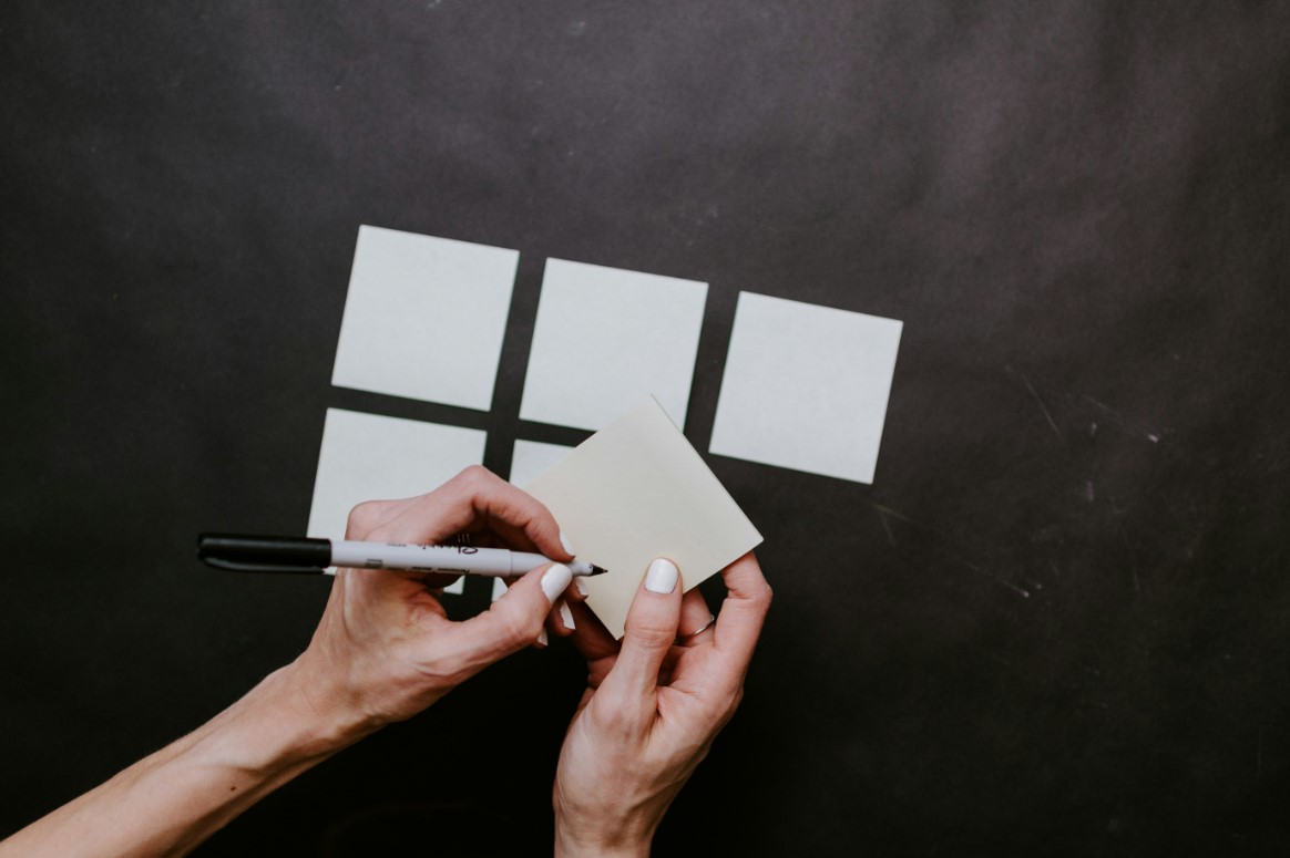 Person writing on a blank sticky note with a marker, with more sticky notes on a black surface.