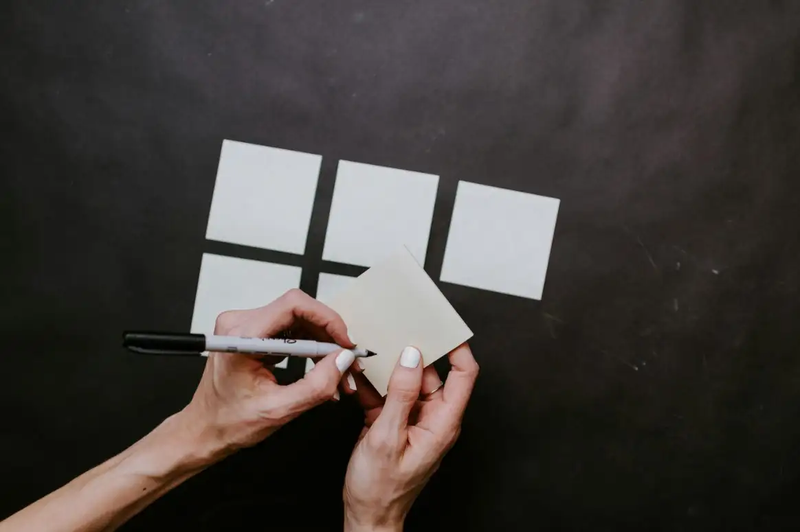 Person writing on a blank sticky note with a marker, with more sticky notes on a black surface.